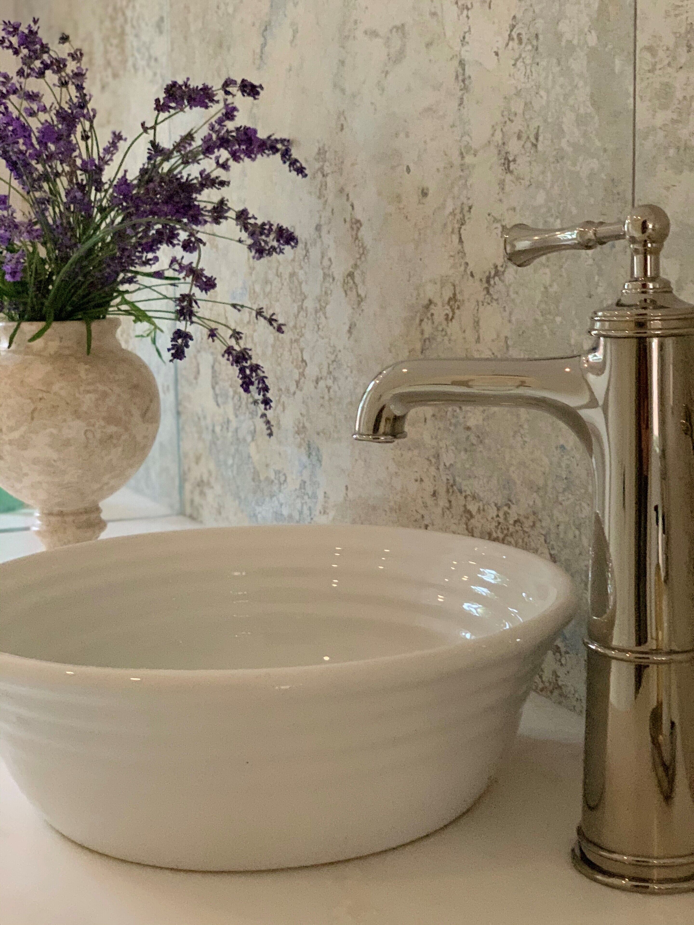 A white ceramic sink with a modern faucet, a vase with purple flowers, and a textured wall in the background.