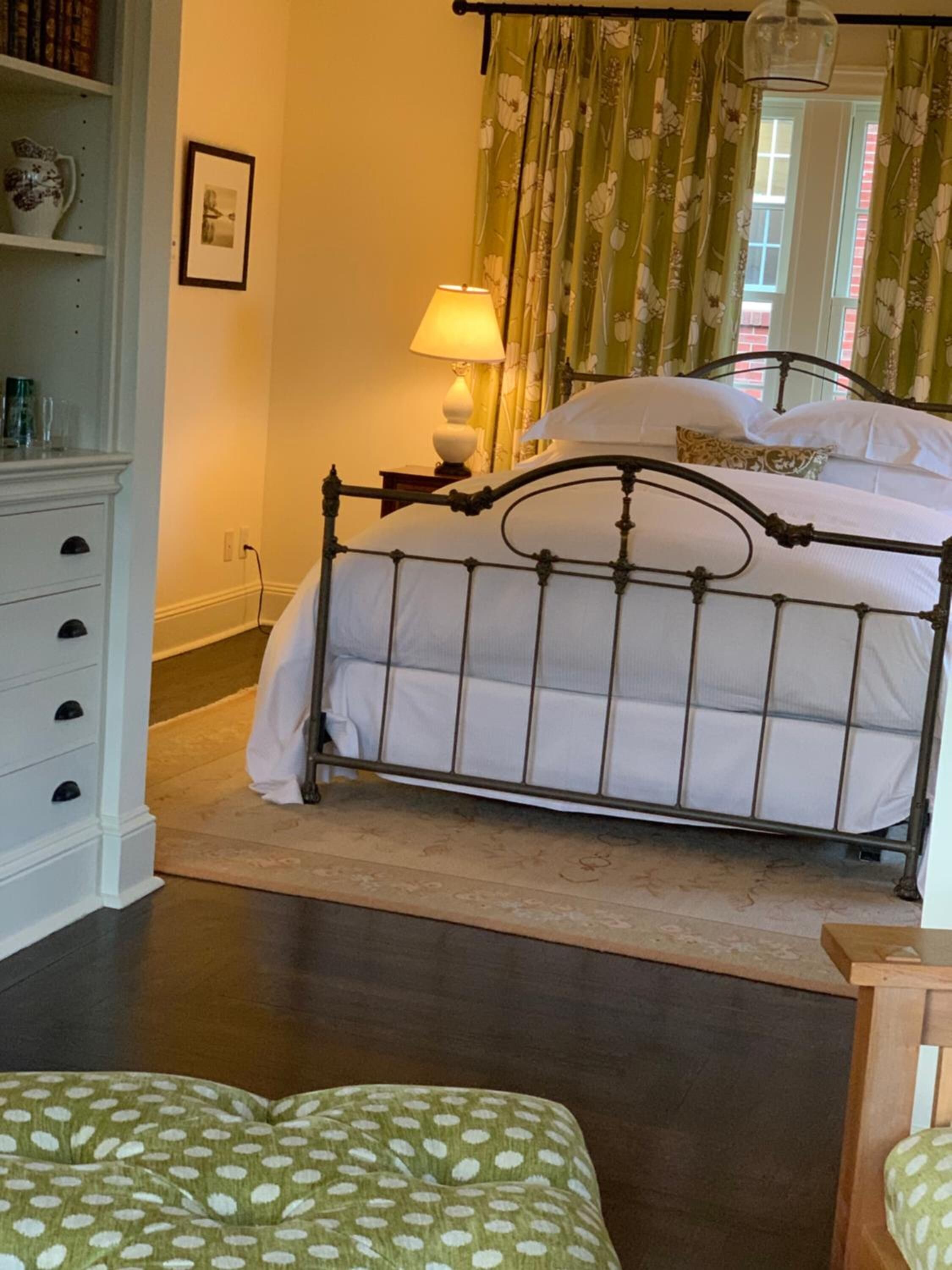 A bedroom with a metal bed frame, a bedside table with a lamp, a dresser, and a green patterned ottoman.