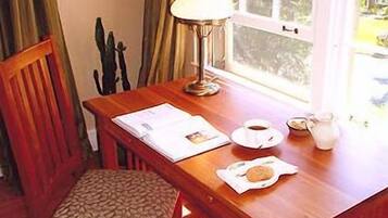 Une table en bois avec une lampe, une chaise, une fenêtre et une table dressée pour le petit-déjeuner, comprenant une tasse, un pichet et une assiette avec une pâtisserie.