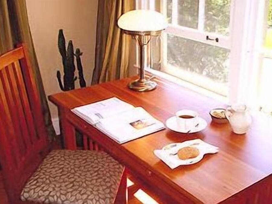 Une table en bois avec une lampe, une chaise, une fenêtre et une table dressée pour le petit-déjeuner, comprenant une tasse, un pichet et une assiette avec une pâtisserie.
