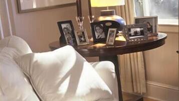 Ein Zimmer mit einem weißen Sofa, einem kleinen runden Tisch mit gerahmten Fotos und einer Lampe, sowie einem Fenster mit Vorhängen.