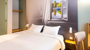 A hotel room with a bed, a nightstand, a chair, and a large window with a view of a canal and buildings. - B&B HOTEL Nantes Parc Expos La Chapelle (La Chapelle-sur-Erdre)