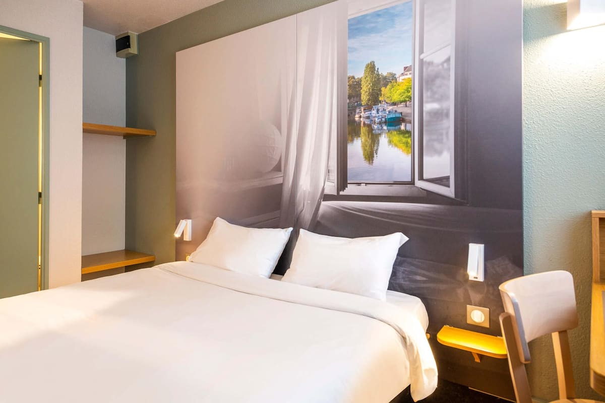 A hotel room with a bed, a nightstand, a chair, and a large window with a view of a canal and buildings.