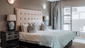 A bedroom with a tufted headboard, two bedside lamps, a nightstand with drawers, a bed with patterned pillows, a light blue blanket, a ceiling fan, and a view of the outside through a large window. - The Mandyville (Jeffreys Bay)