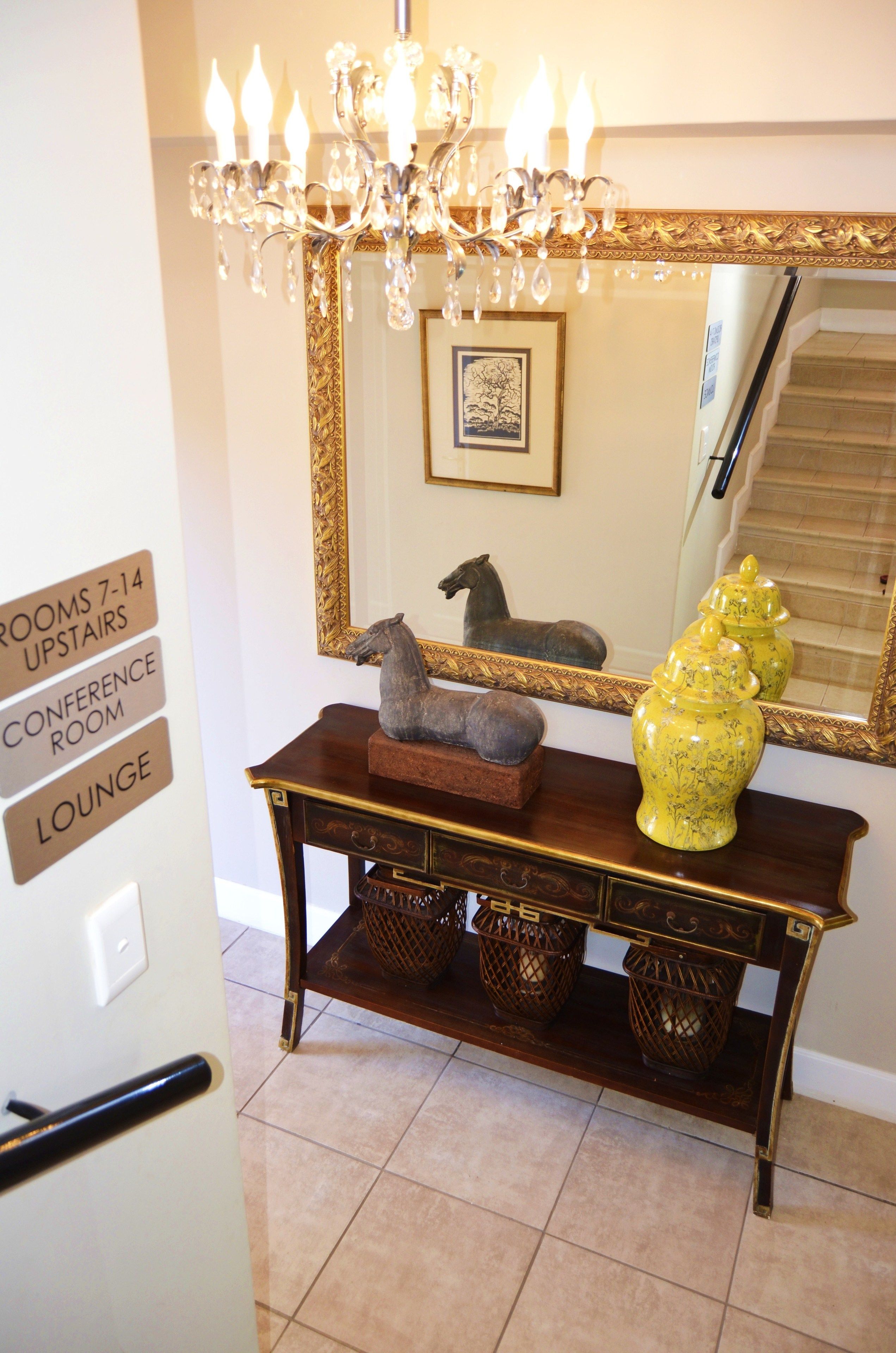 A lobby with a chandelier, a console table with decorative items, a mirror reflecting a staircase, and a sign indicating room locations.