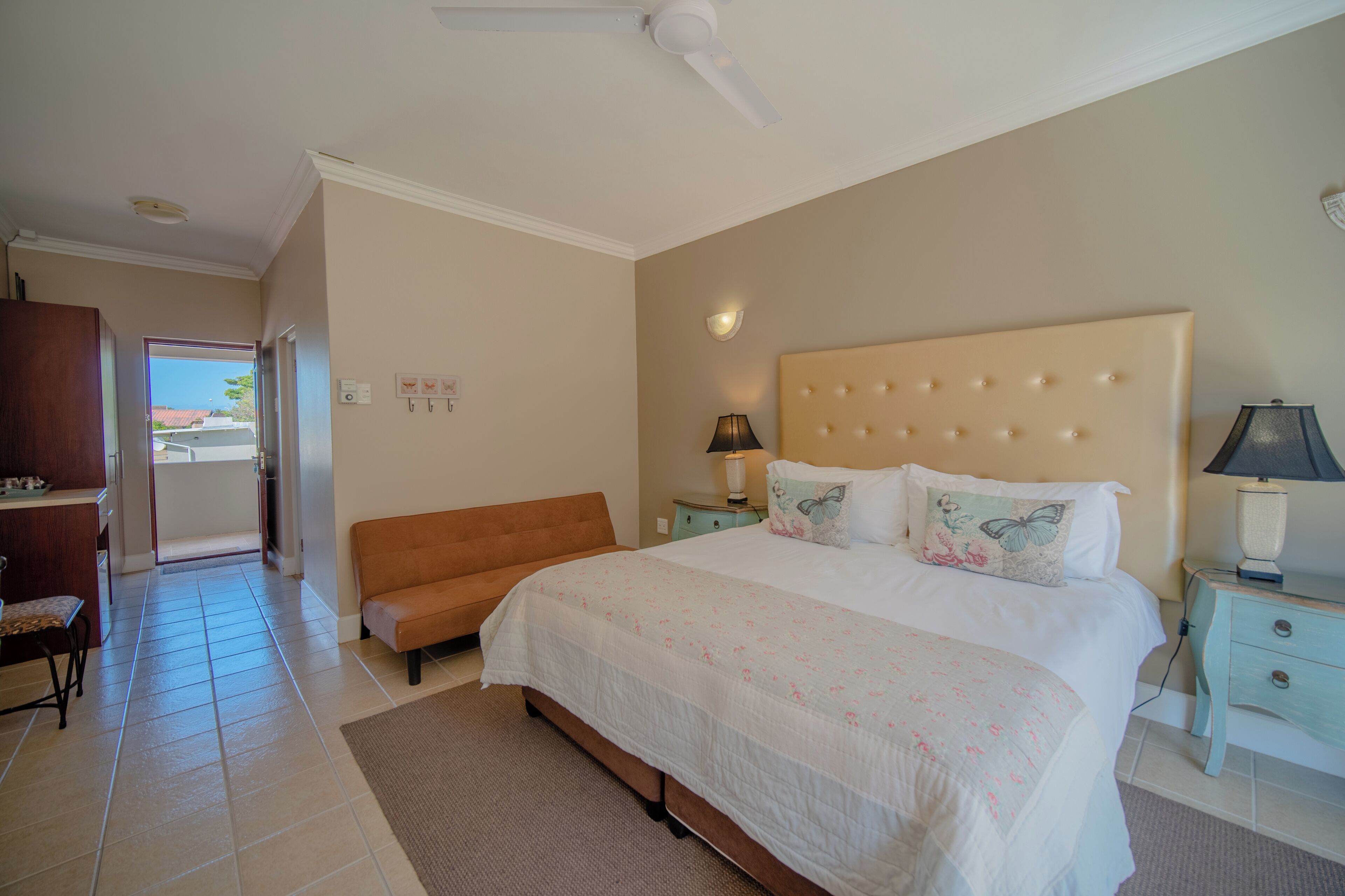 A bedroom with a large bed, a nightstand with a lamp, a sofa, and a ceiling fan.