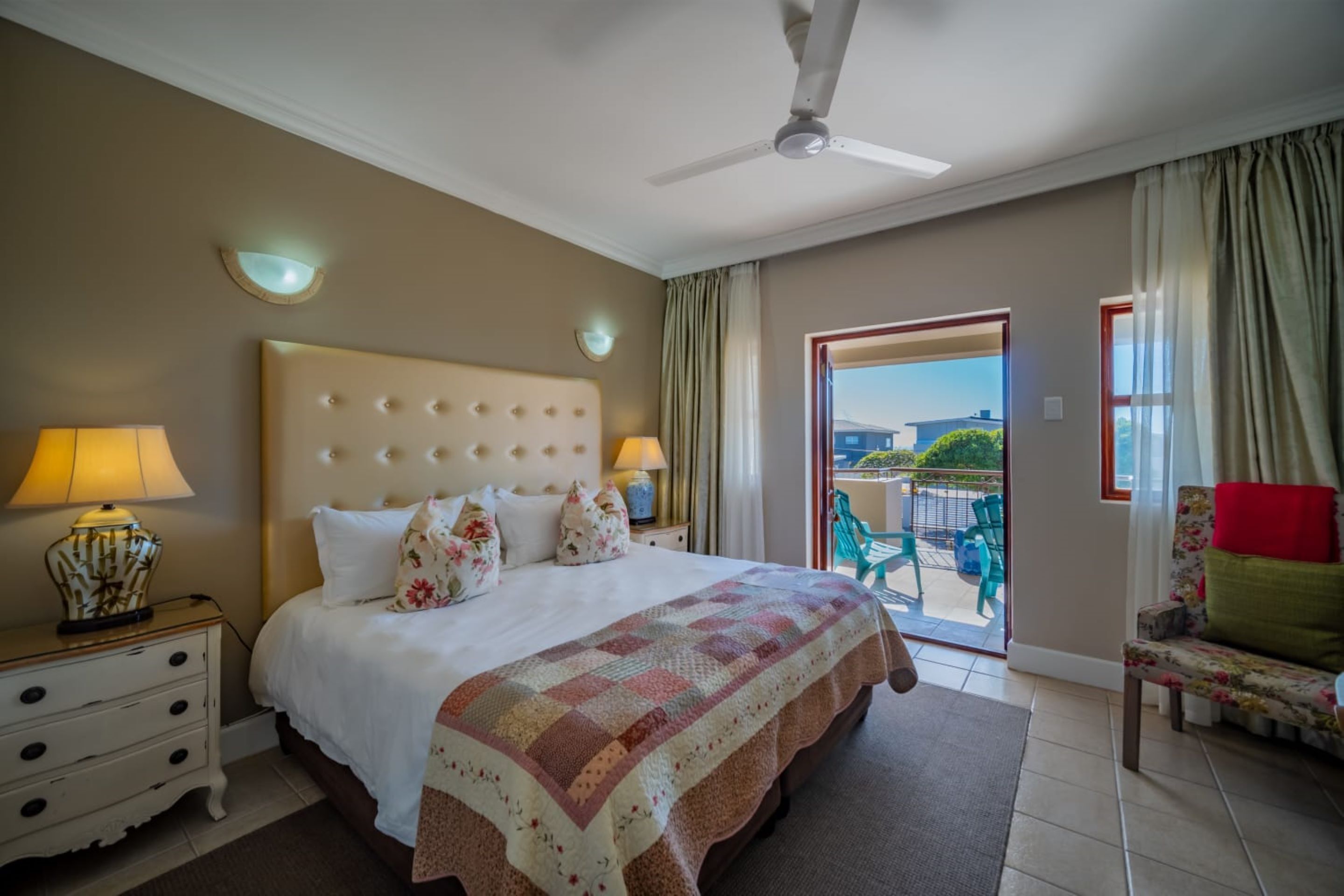 A hotel room with a large bed, two bedside lamps, a ceiling fan, a view to the outside, and a chair by the window.