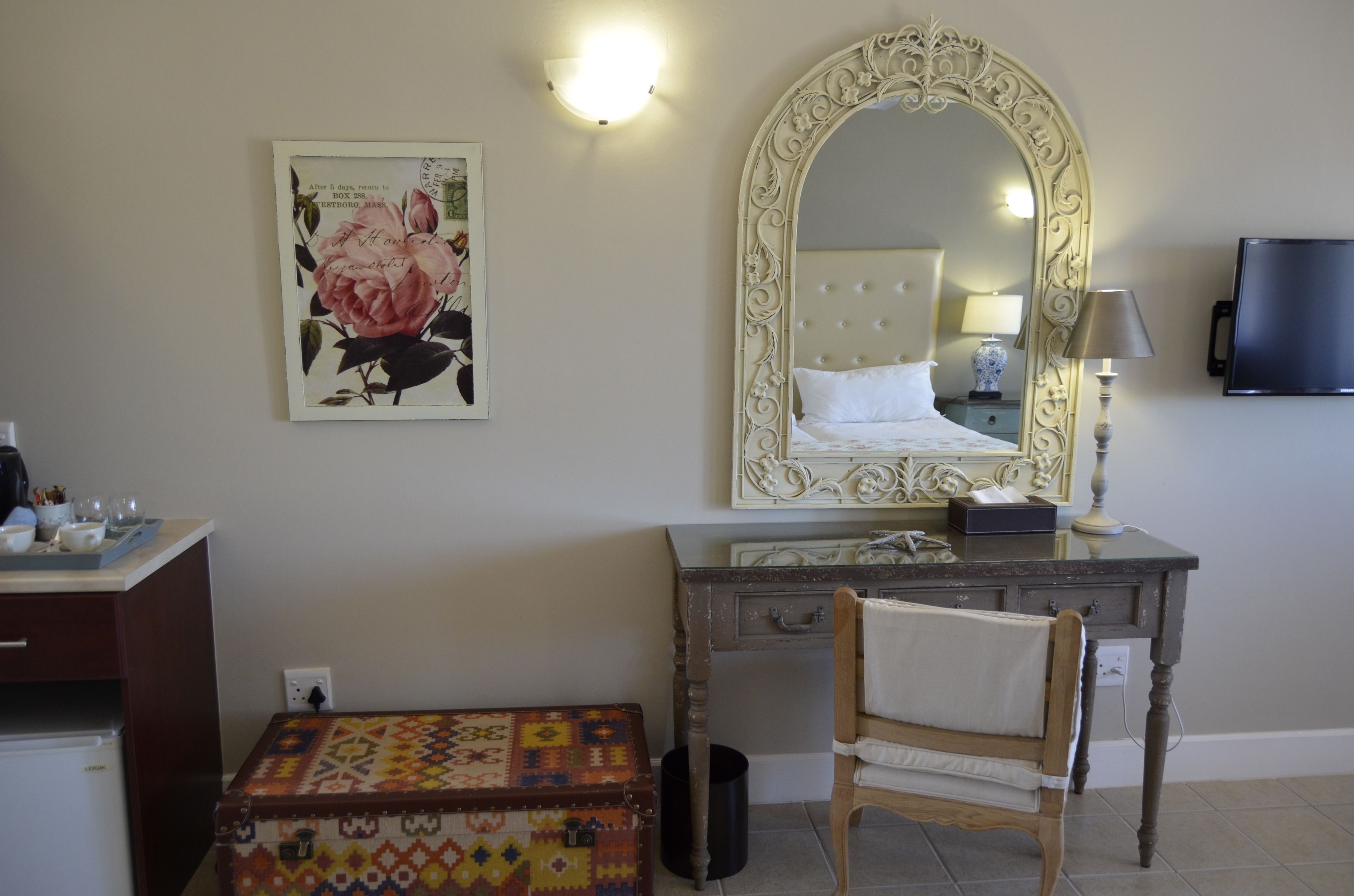 A bedroom with a large ornate mirror, a bedside table with a lamp, a wooden chair, a small table with a decorative box, and a painting on the wall.