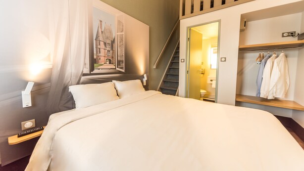 Habitación de hotel con una cama grande, un cuadro de un edificio, un armario con ropa colgada y una escalera.