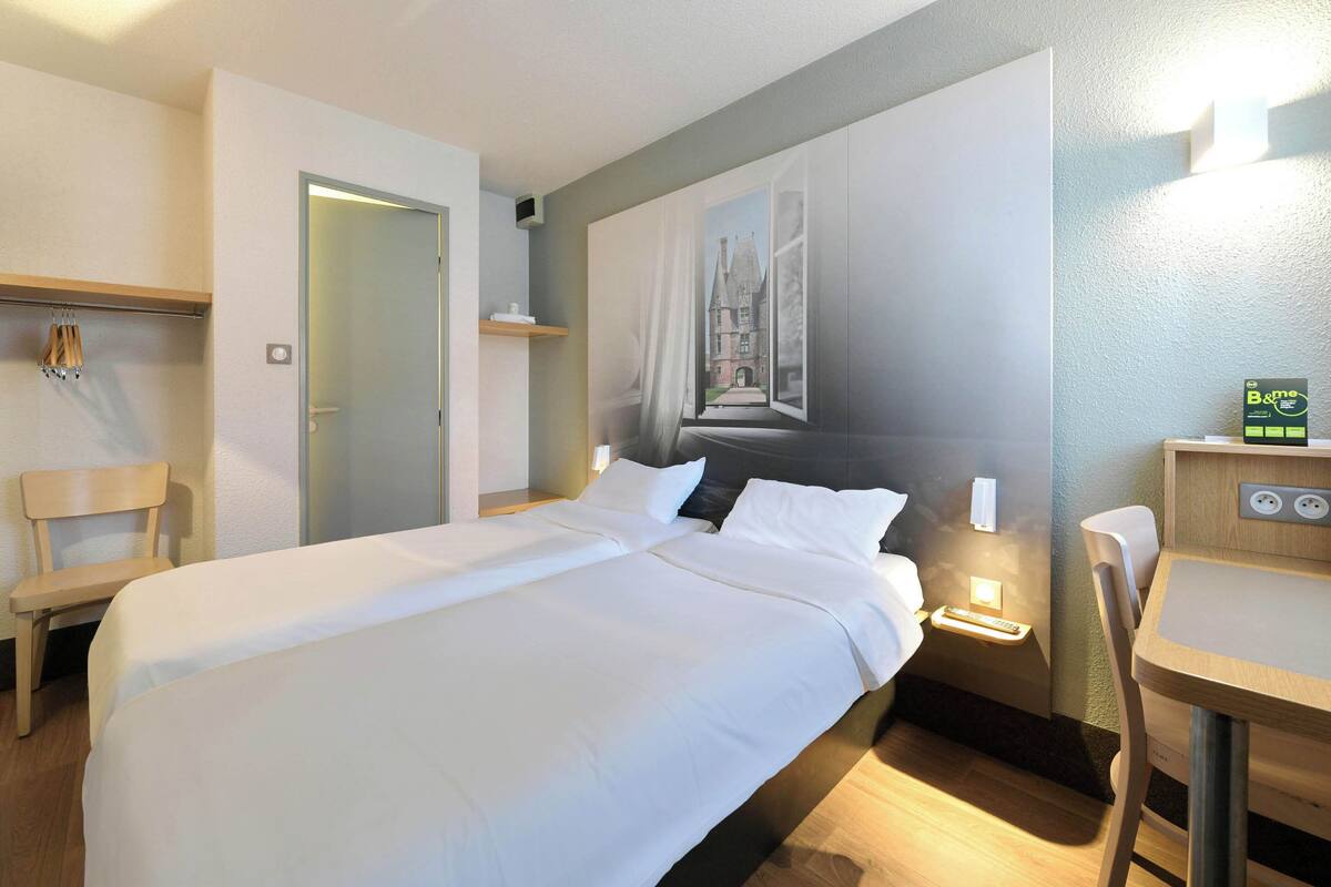 A neatly made bed with white linens, a bedside table with a lamp, a chair, and a wall-mounted shelf.