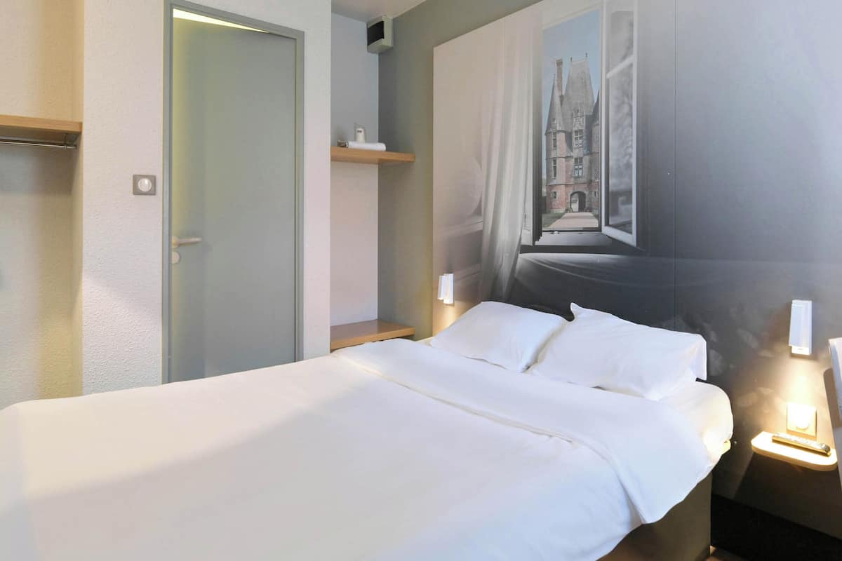 A hotel room with a bed, a nightstand, a wall-mounted shelf, and a door.