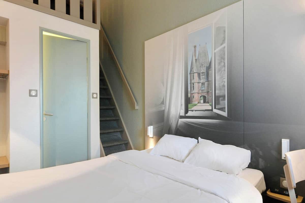 A hotel room with a bed, a staircase, and a large window with a view of a building.