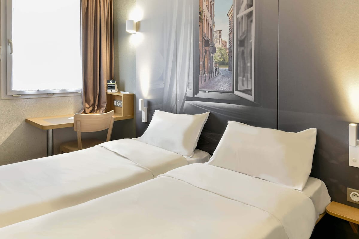 A hotel room with two beds, a desk, a chair, and a view of buildings through a window.
