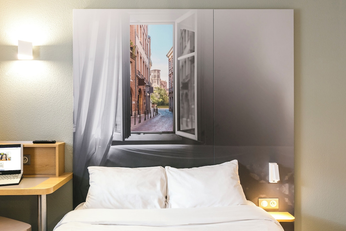 A hotel room with a bed, a desk with a laptop, a nightstand with a lamp, and a large window with a view of buildings.