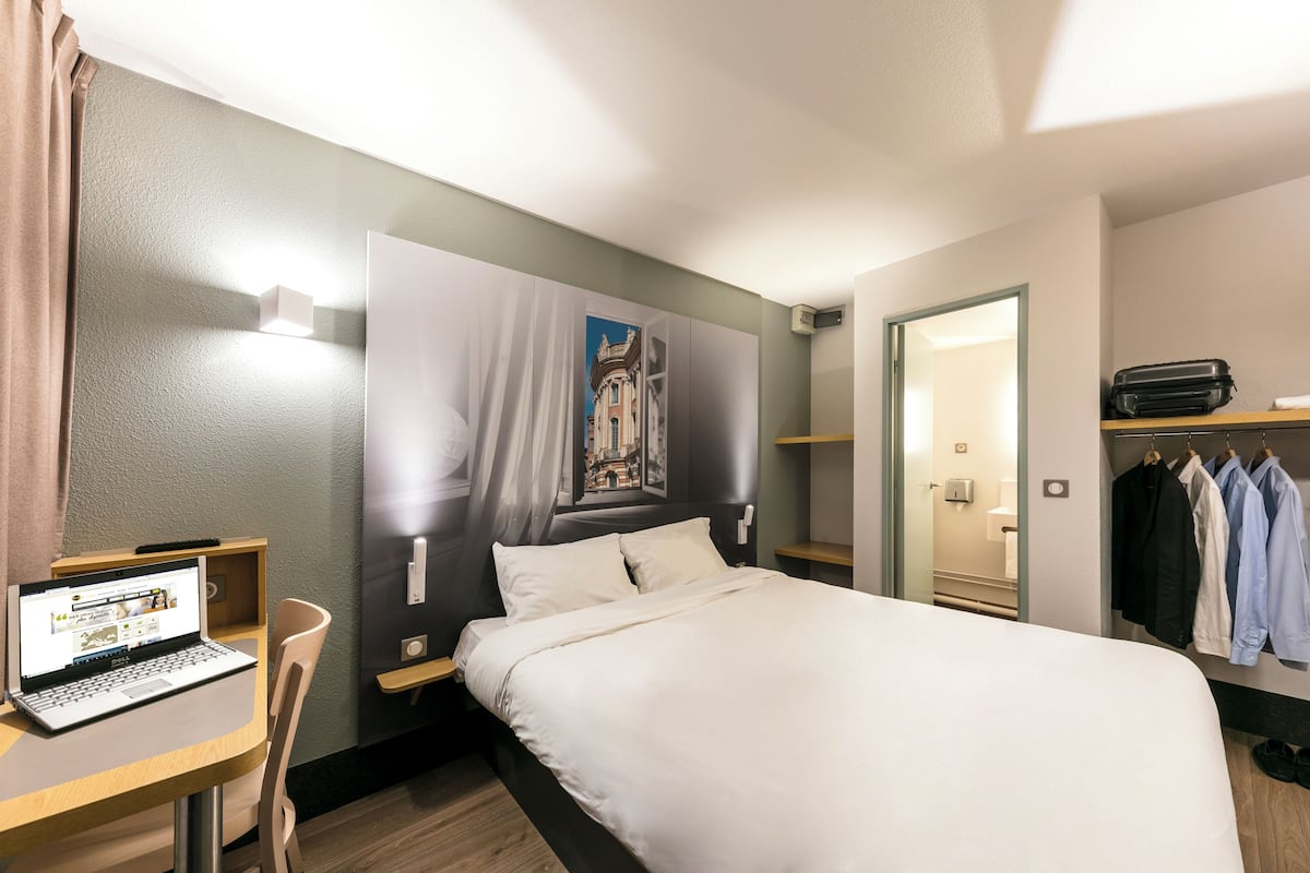 A hotel room with a bed, a desk with a laptop, a chair, a wardrobe, and a painting on the wall.