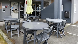 An outdoor seating area with tables and chairs, a yellow umbrella, and a building entrance with a red awning. - B&B HOTEL Toulouse Cité de l'Espace Mouchotte (Toulouse)