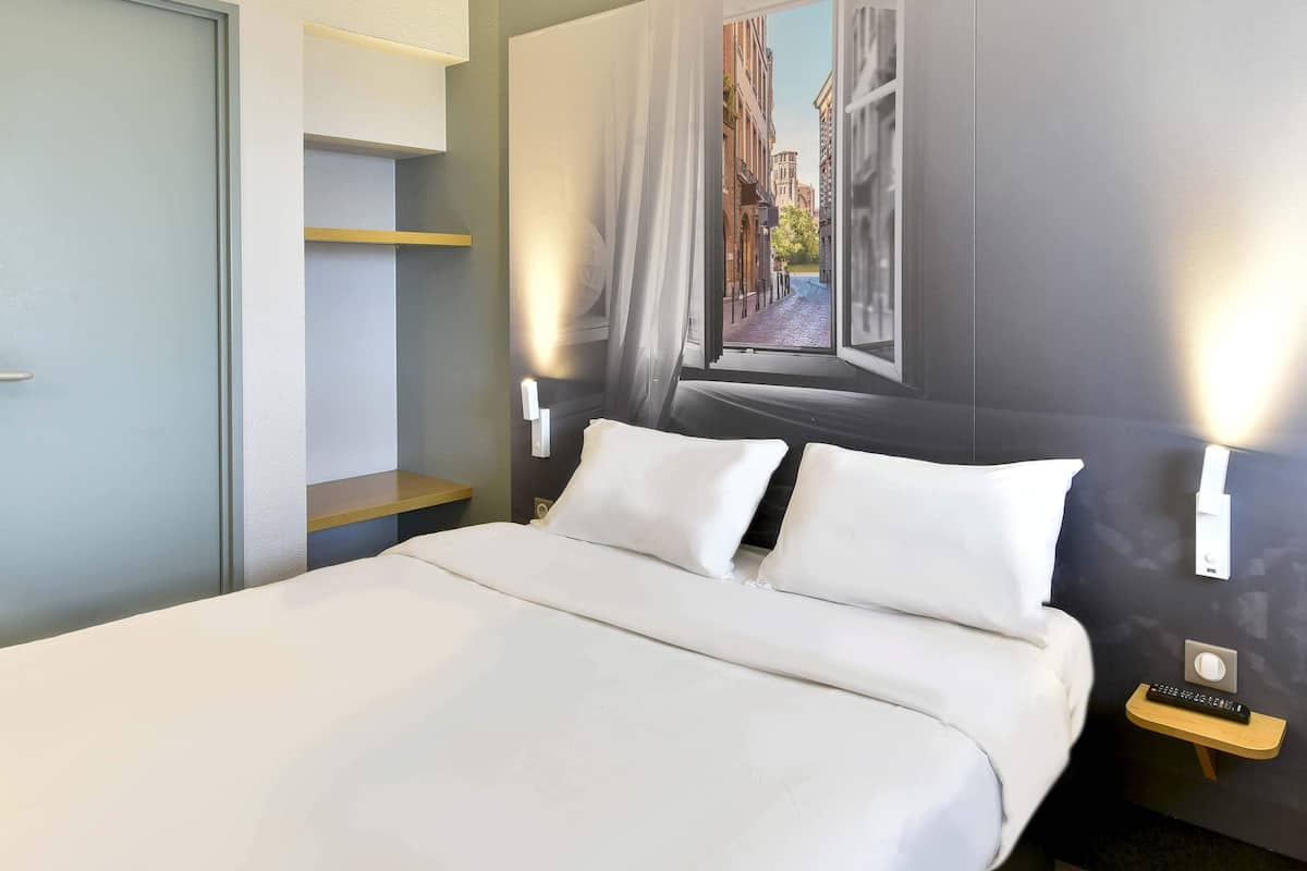 A hotel room with a bed, a nightstand, a wall-mounted TV, and a window with a view of buildings.