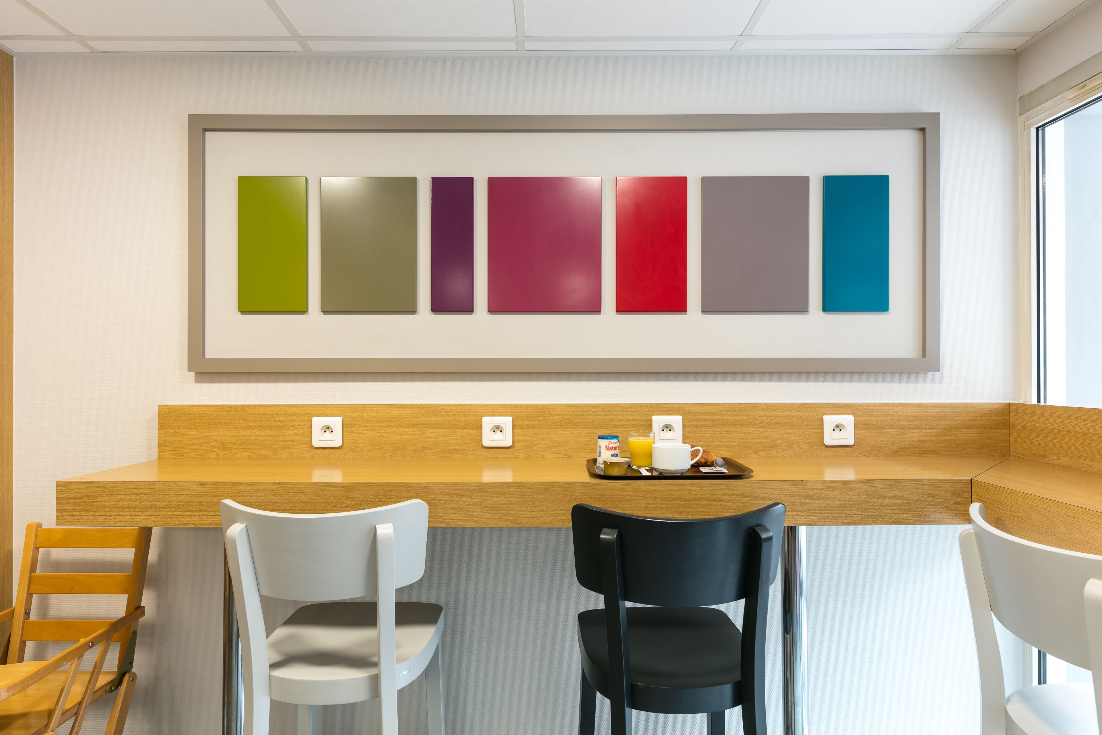 Un espace moderne dédié au petit-déjeuner, avec un comptoir en bois, des chaises blanches et noires, et un mur présentant différents échantillons de couleurs.
