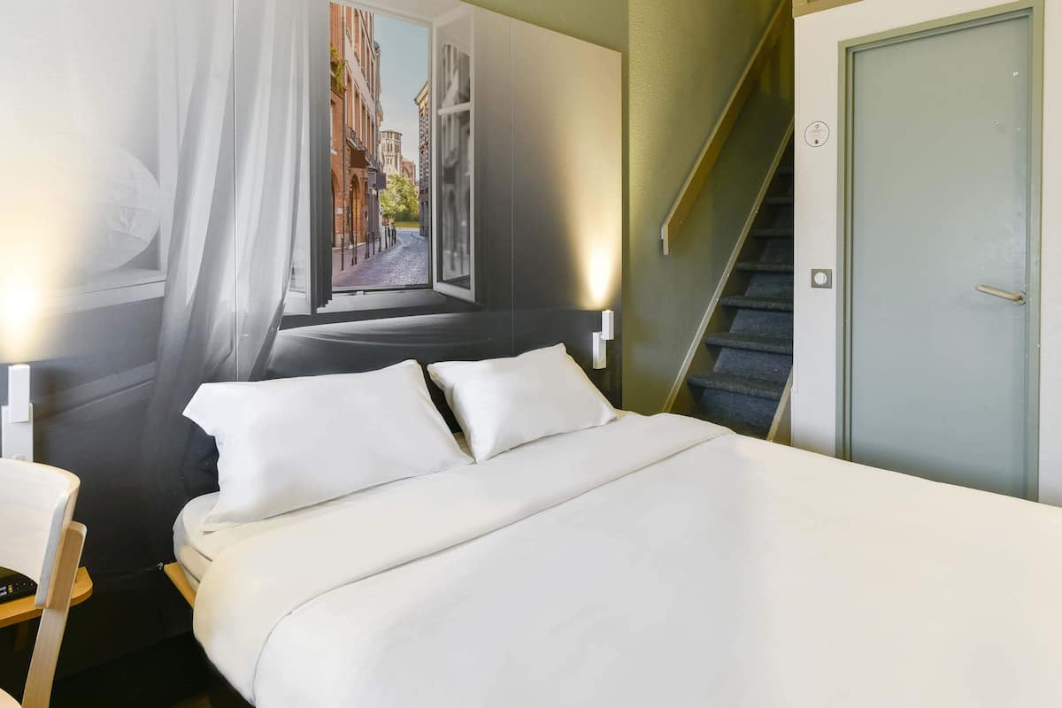 A hotel room with a bed, a chair, a desk, a window with a view of buildings, and a door with a staircase.