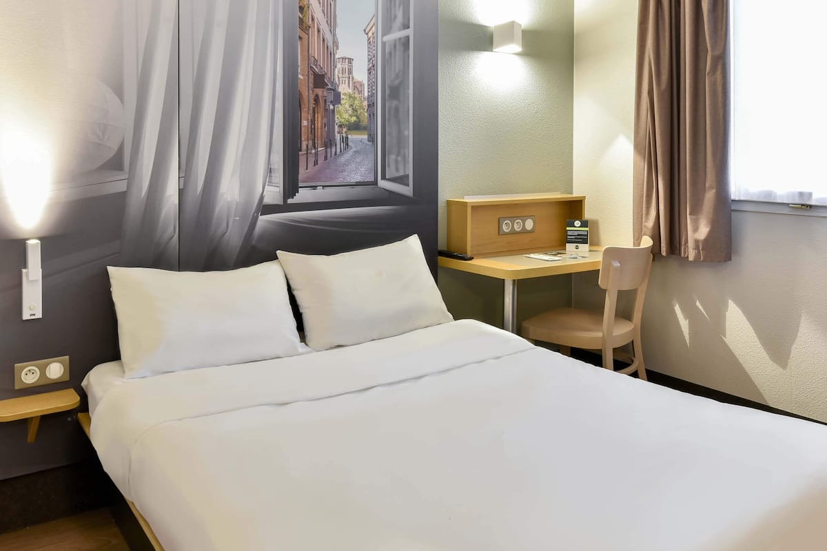 A hotel room with a bed, a desk with a chair, a wall-mounted TV, and a window with curtains.