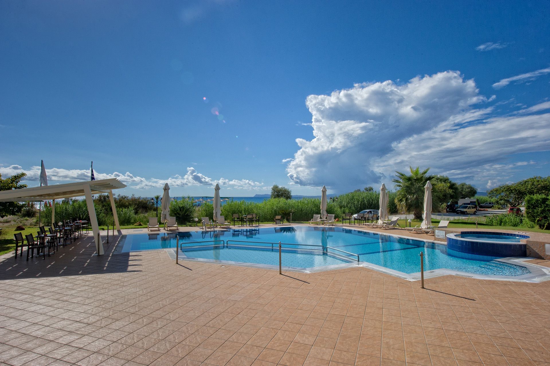 Una piscina con sillas de descanso y sombrillas, rodeada de follaje y un cielo despejado.