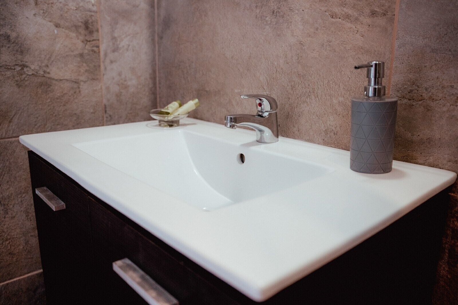A bathroom with a white sink, a modern faucet, a soap dispenser, and a small glass container.