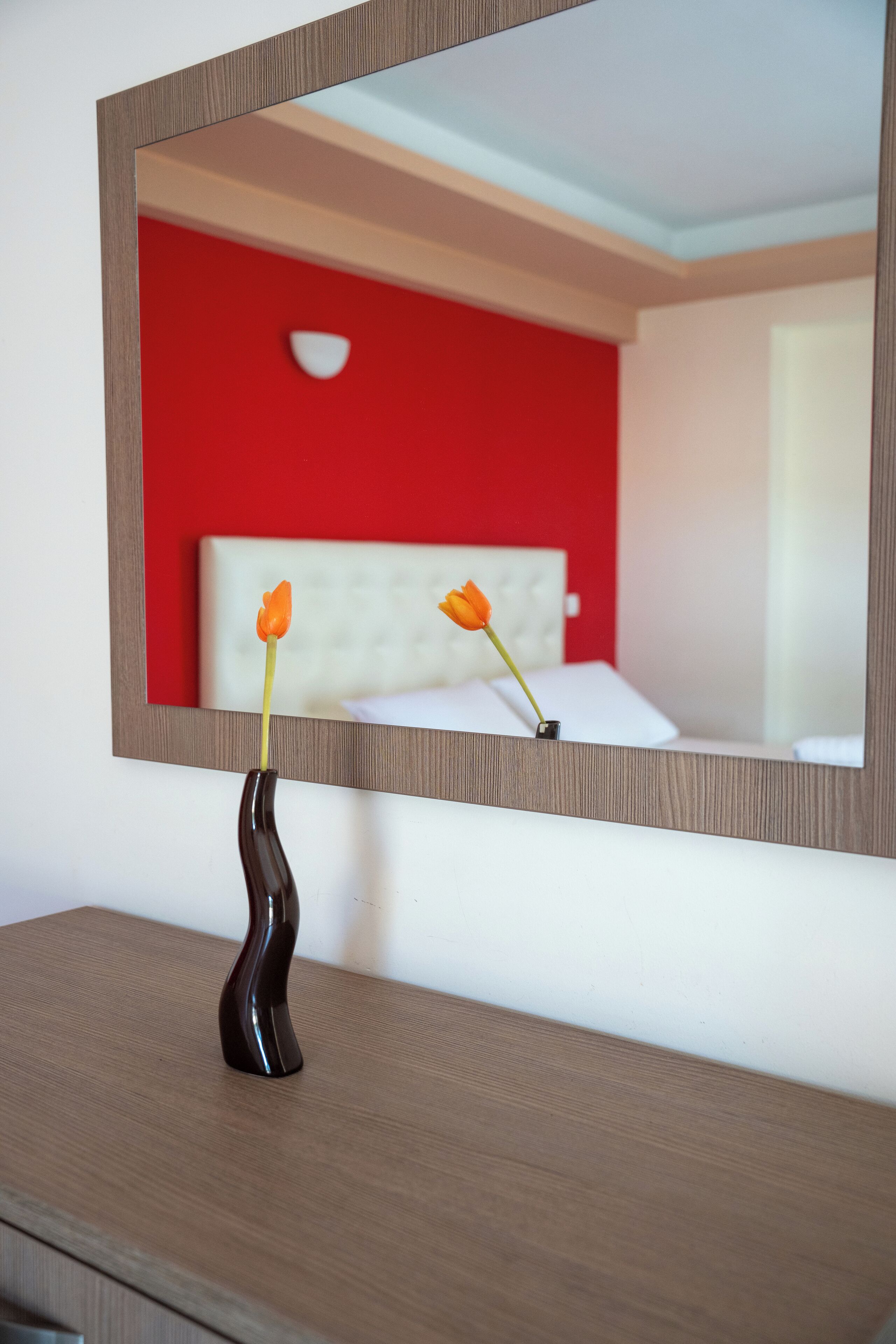 A mirror reflecting a room with a red wall, a white headboard, and a vase with two orange flowers on a wooden dresser.