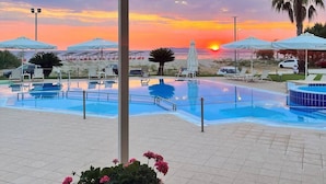 Zona de piscina con sombrillas y sillas de descanso, vista al atardecer y una palmera.