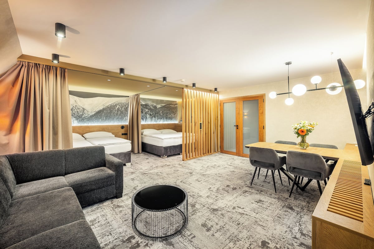 A modern hotel room with a sofa, a round coffee table, a bed with a mountain view, a dining area with a table and chairs, and a TV.