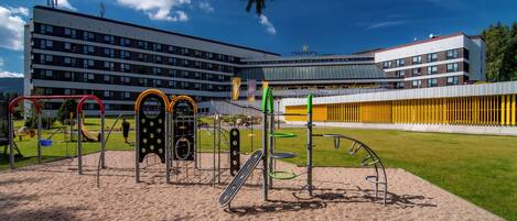 Un parque infantil con diversas estructuras de juego, una zona verde y un edificio con múltiples ventanales al fondo.