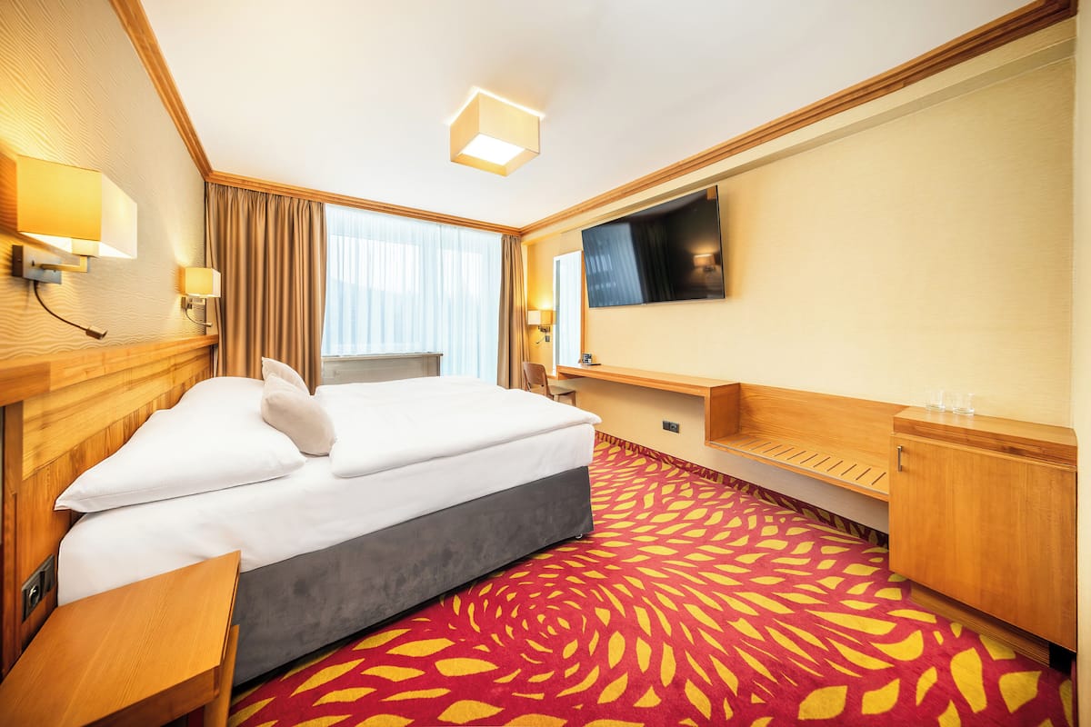 A hotel room with a large bed, a TV, a desk, and a red patterned carpet.