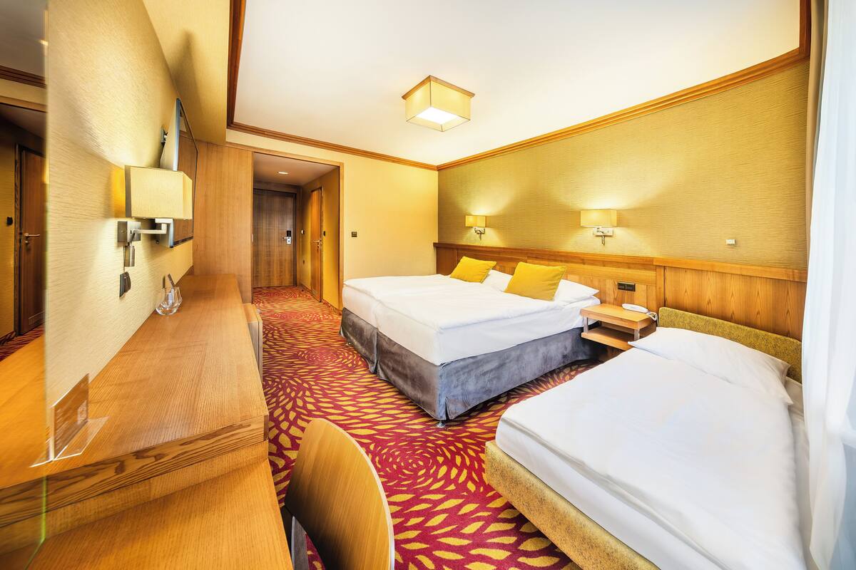 A hotel room with two beds, a desk, and a chair. There are wall-mounted lamps and a door leading to another room.
