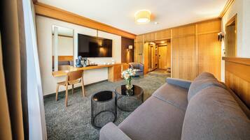 Ett modernt hotellrum med en grå soffa, ett skrivbord i trä, en platt-TV och ett litet soffbord med en vas med blommor.