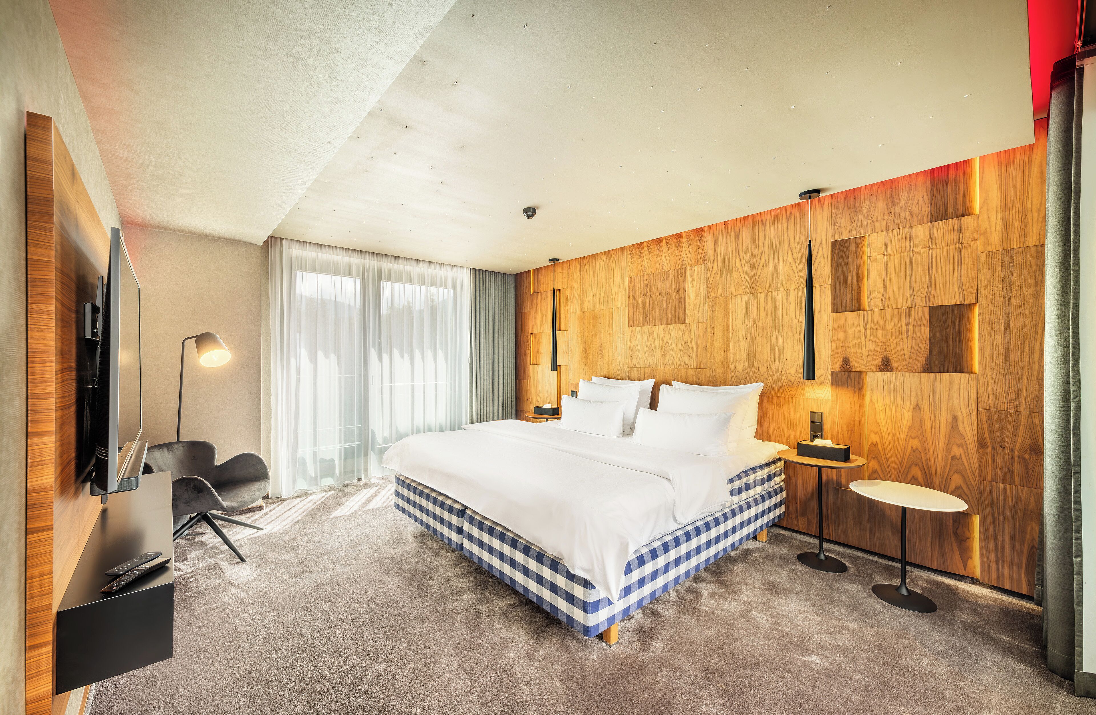 A modern hotel room with a large bed, a desk with a chair, a TV, and a small table with a lamp.