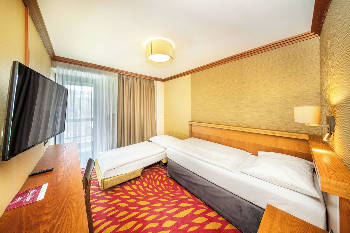 A hotel room with a TV, two single beds, a wooden headboard, a red patterned carpet, and a window with curtains.