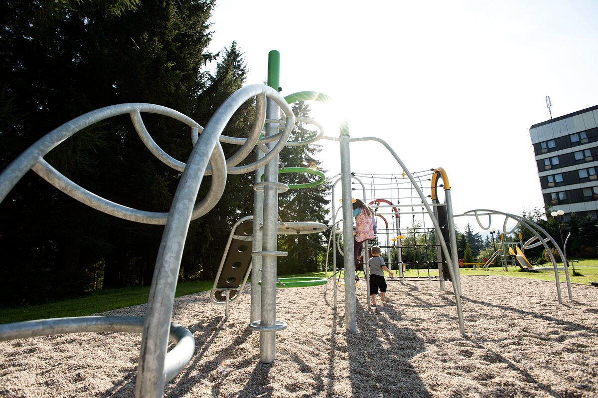 A playground with metal climbing structures and a slide, surrounded by trees and a building in the background.