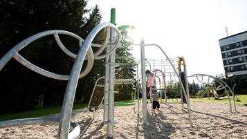 A playground with metal climbing structures and a slide, surrounded by trees and a building in the background.