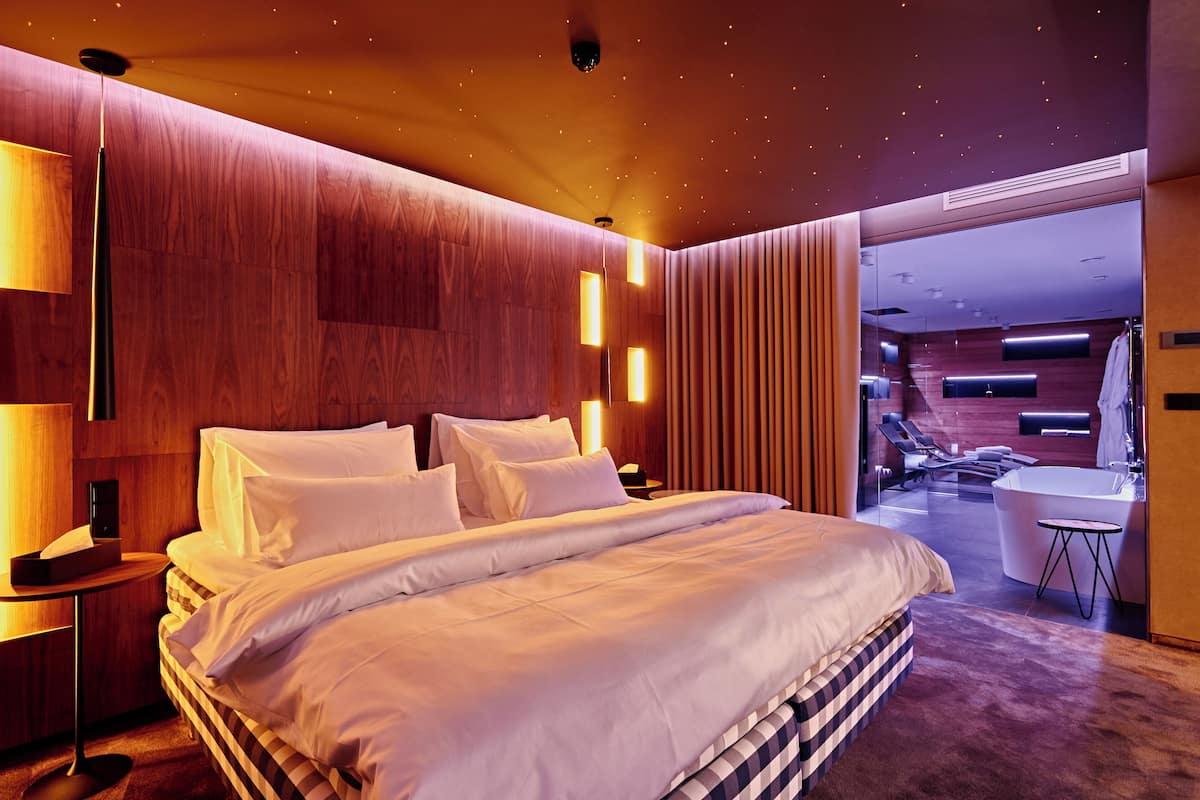 A modern hotel room with a large bed, bedside lamps, a vanity with a mirror, and a view into the bathroom.