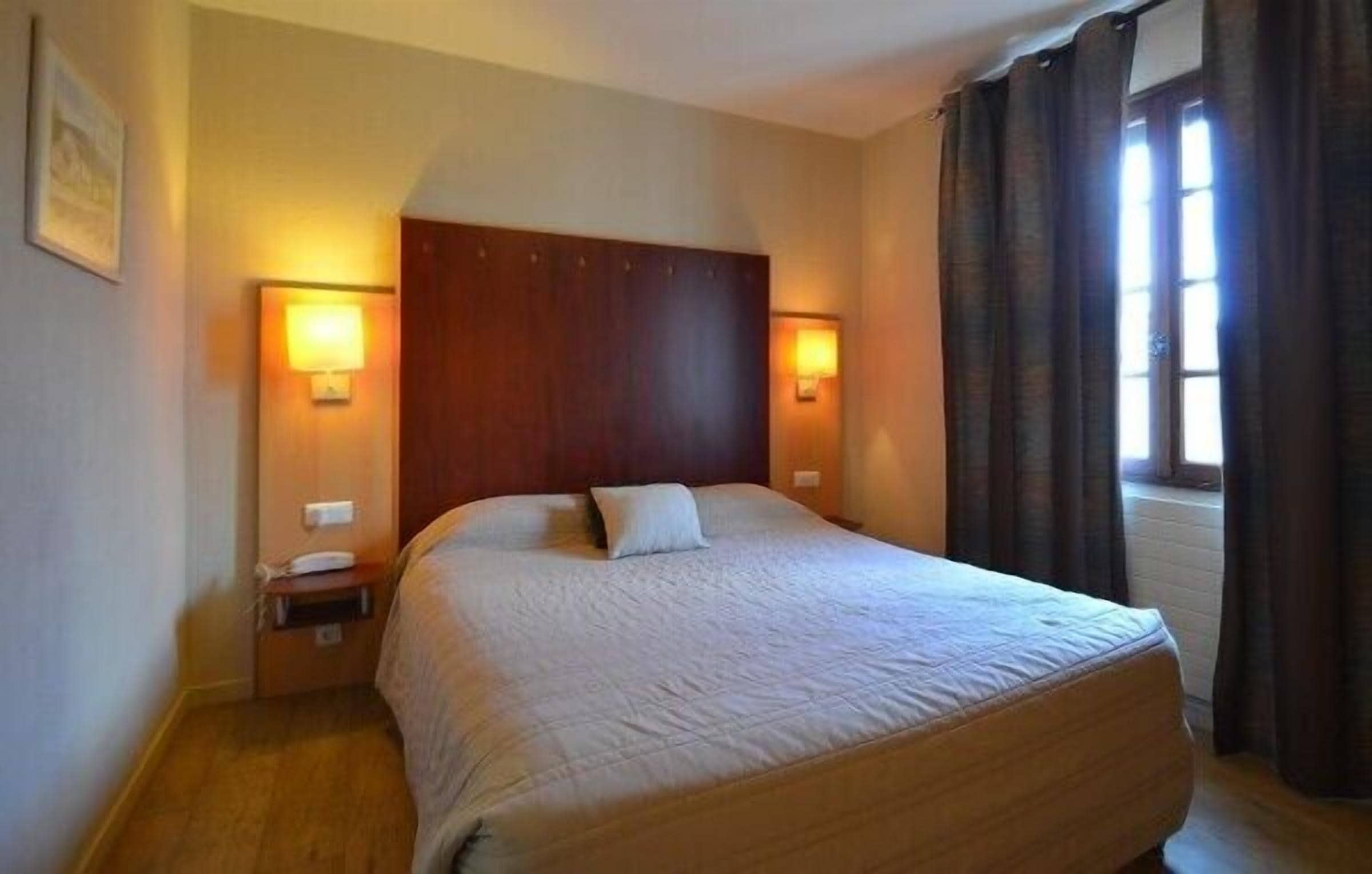 Una habitación de hotel con una cama grande, dos lámparas de noche, una mesita de noche y una ventana con cortinas.