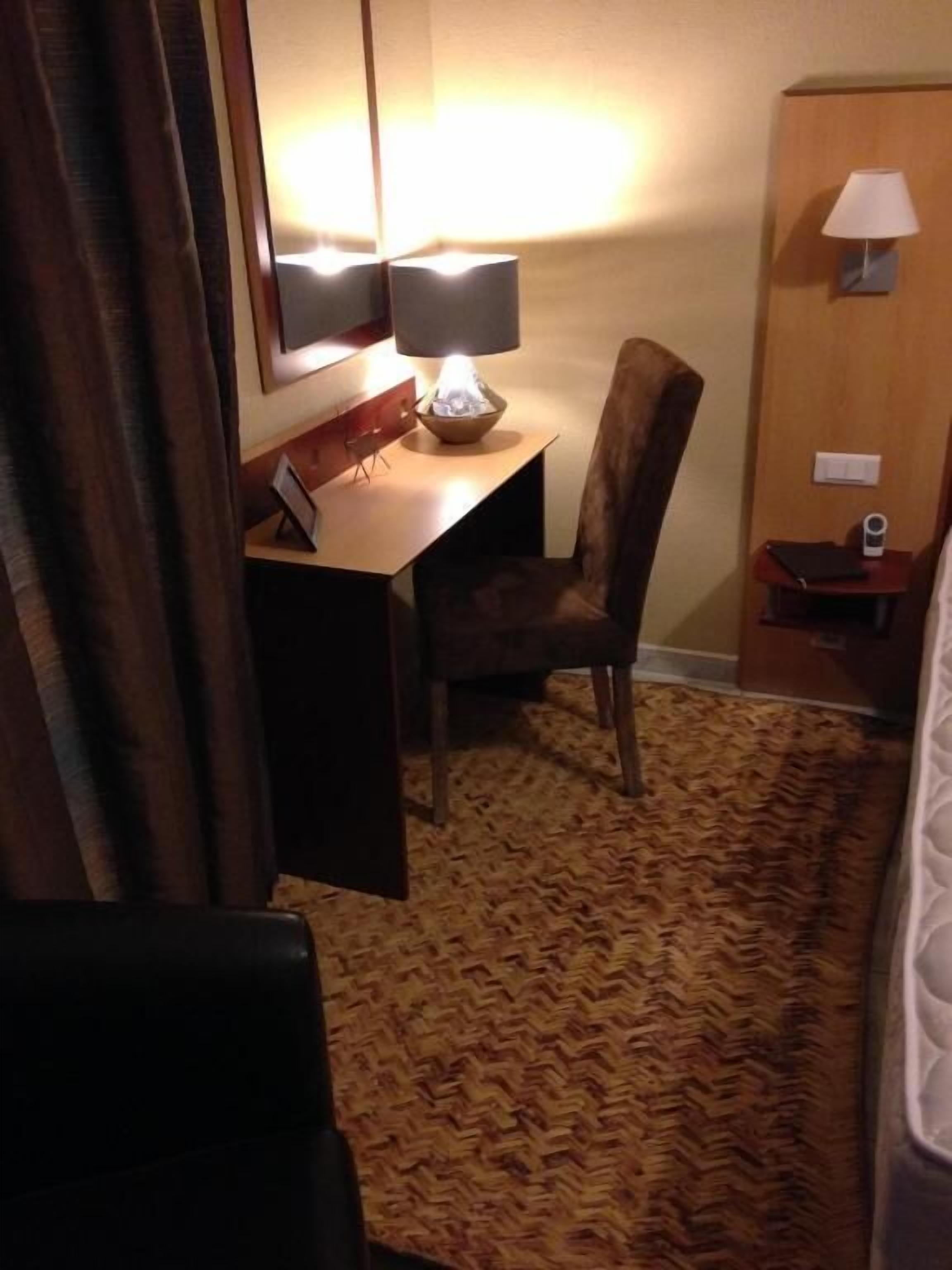 A hotel room with a desk, chair, lamp, and a bed.