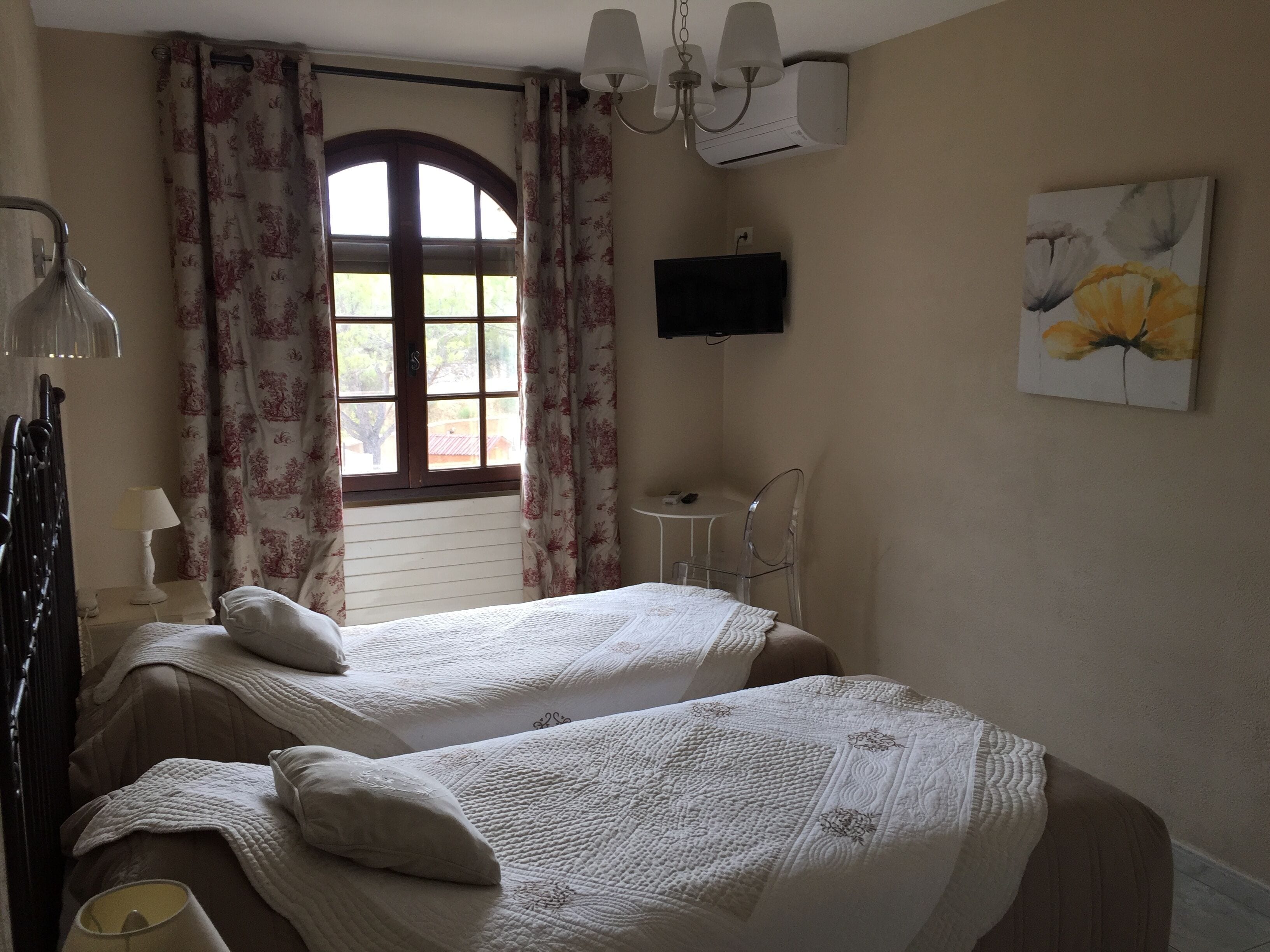 Quarto com duas camas, uma escrivaninha, uma cadeira, uma TV e uma janela com cortinas.