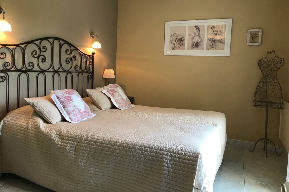 A neatly made bed with a quilted cover and decorative pillows, a wall-mounted picture frame with sketches, and a mannequin dress stand.