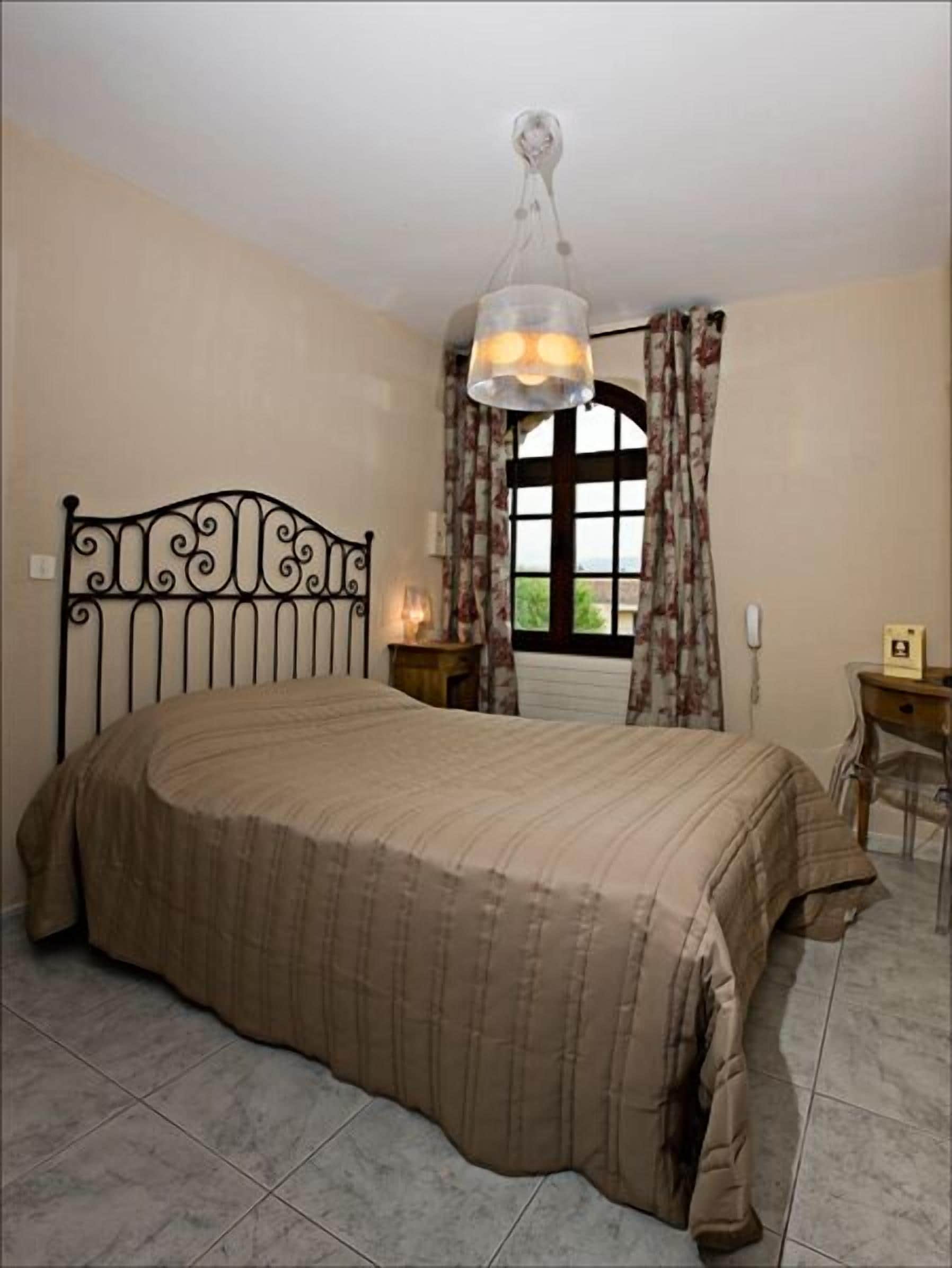 A bedroom with a bed, a desk, a chair, a window with curtains, and a hanging light fixture.