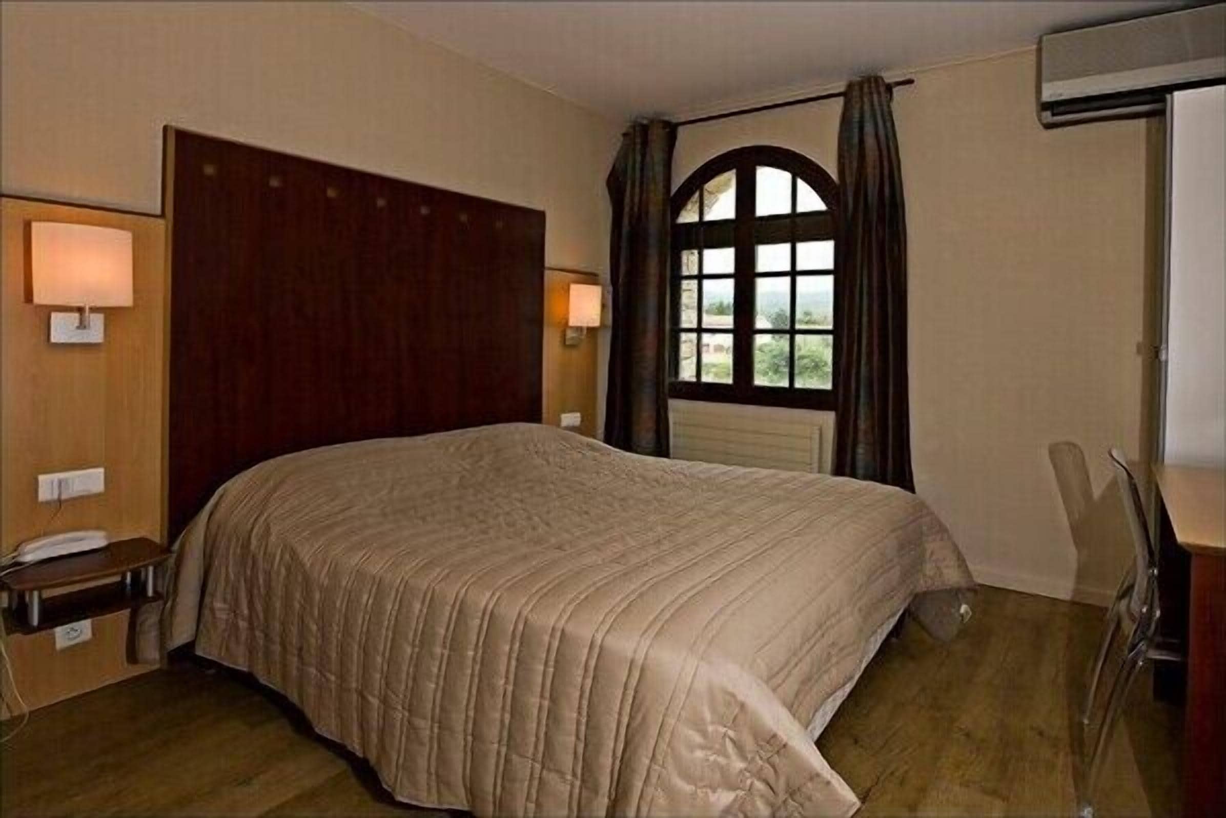 Ein Hotelzimmer mit einem großen Bett, einem hölzernen Kopfteil, einem Fenster mit Vorhängen, einem Schreibtisch mit Lampe und einem Sessel.
