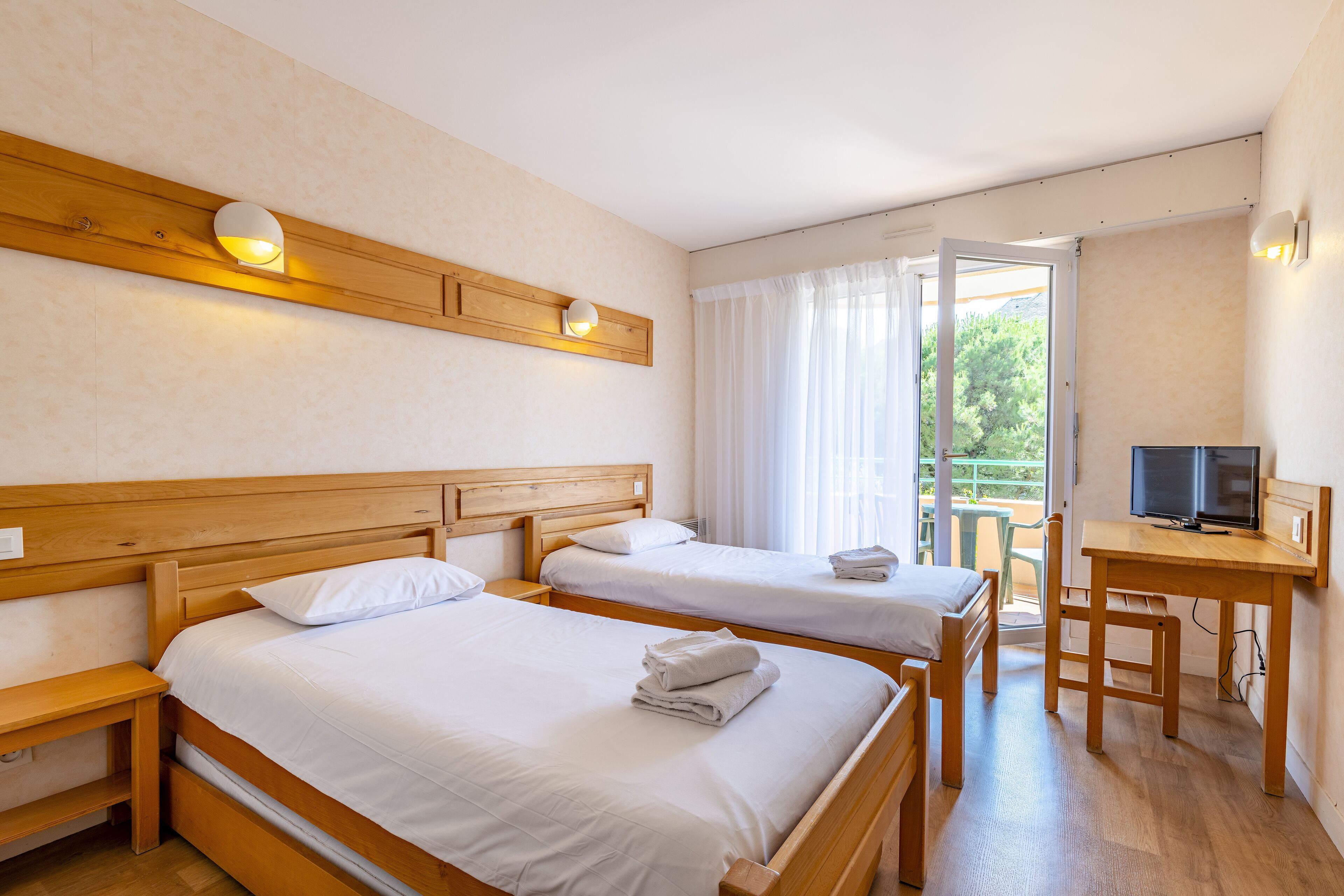 A hotel room with two single beds, a wooden desk, a television, and a balcony with a view of greenery.