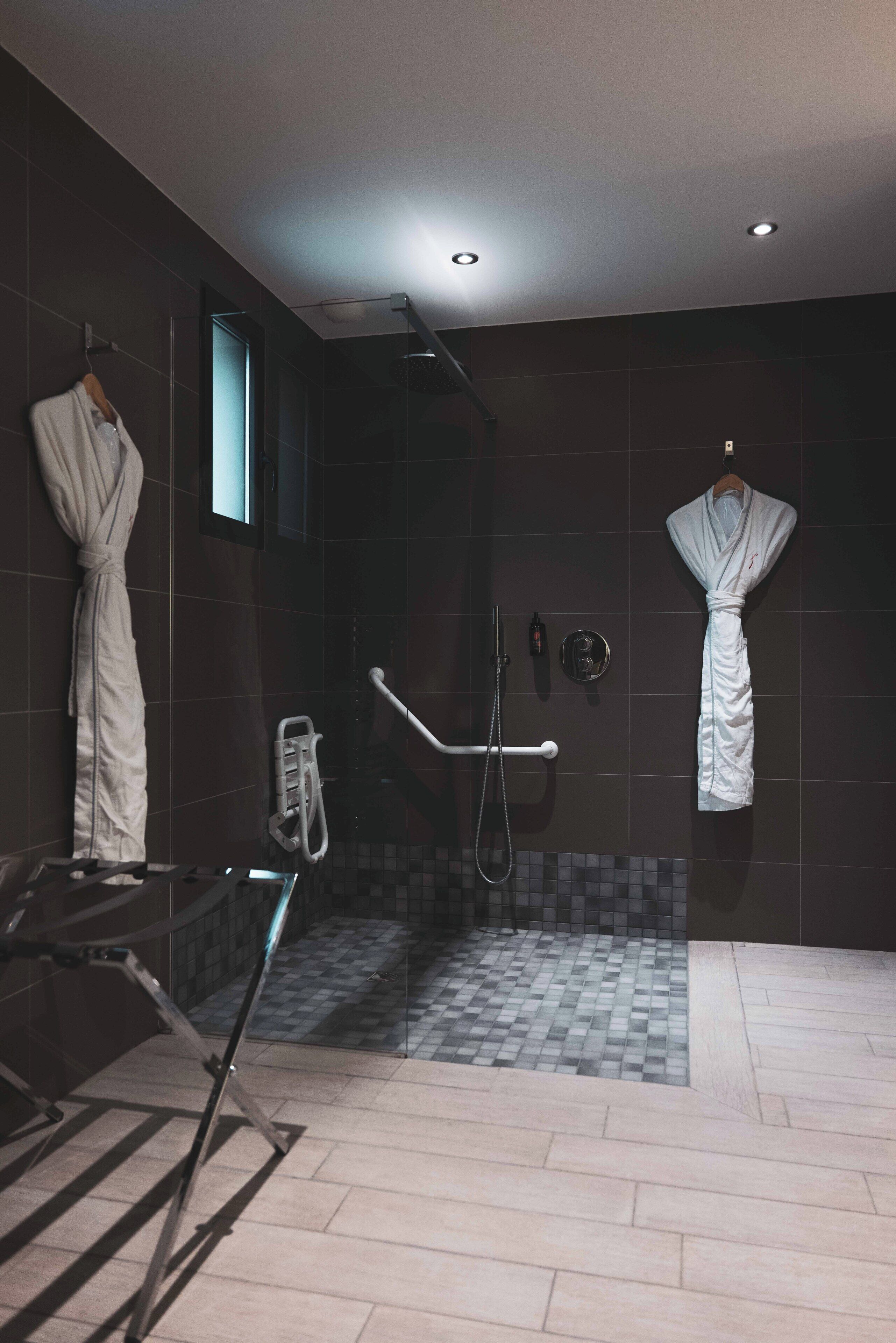 Un bagno moderno con piastrelle scure, cabina doccia in vetro e due accappatoi bianchi appesi a ganci.