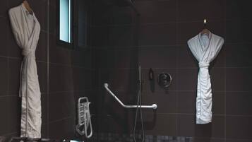 Un bagno moderno con piastrelle scure, cabina doccia in vetro e due accappatoi bianchi appesi a ganci.