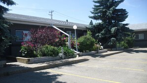 A parking lot with a building that has a sloped roof and a small garden with pink flowers. - Stardust Motel (Camrose)