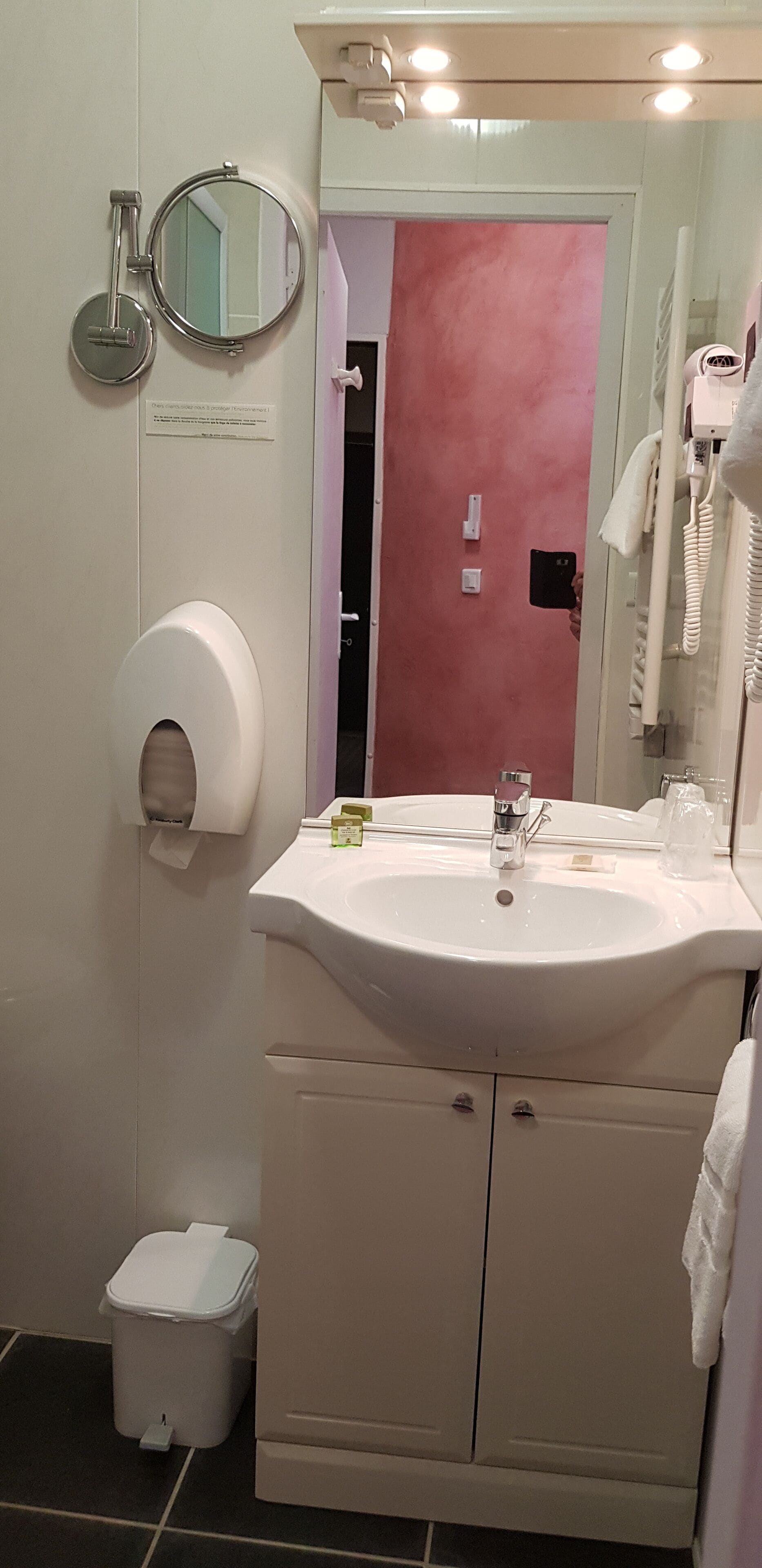 A bathroom with a white sink, a mirror, a toilet paper dispenser, a trash bin, and a towel hanging on the wall.