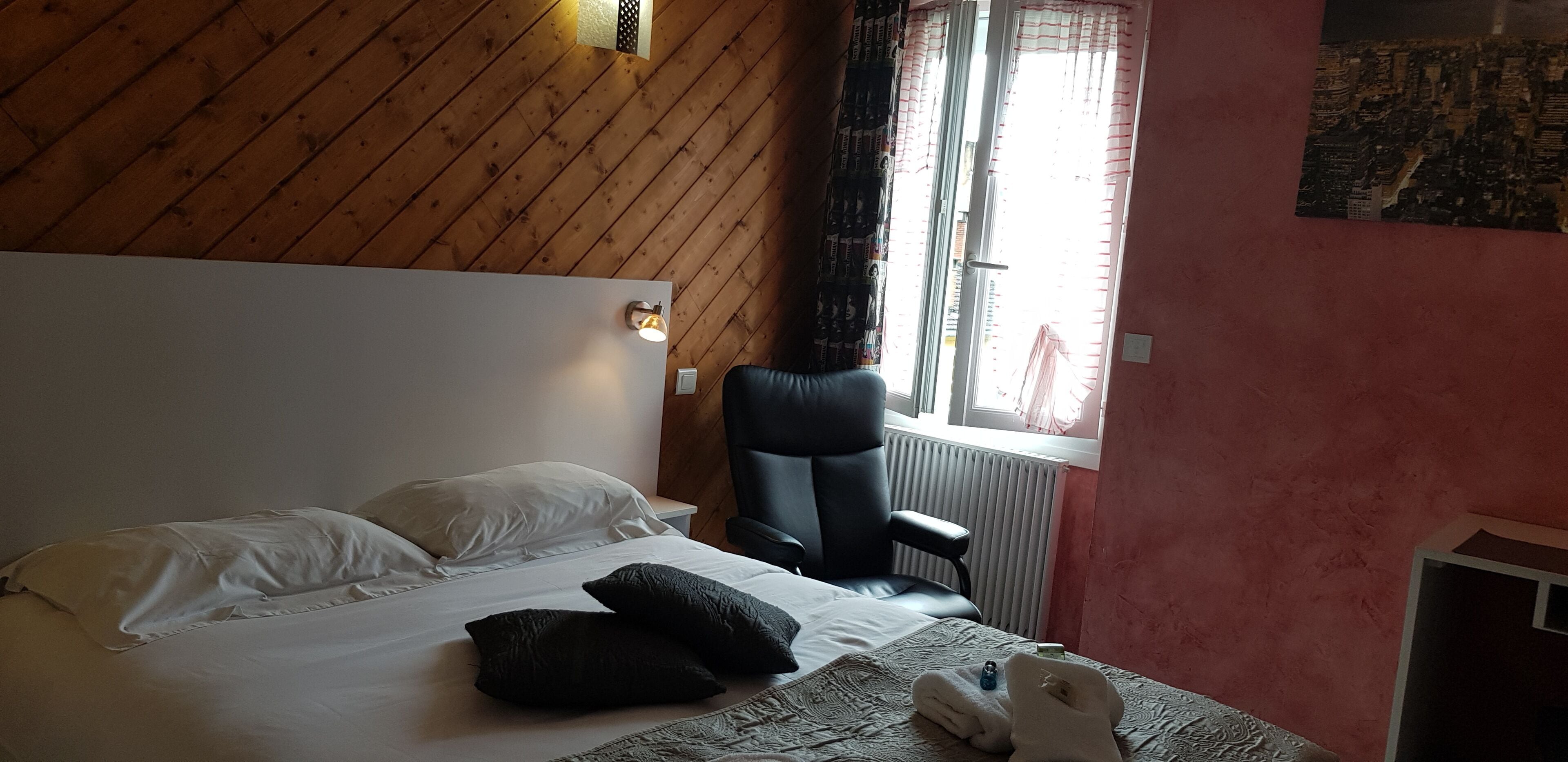 Habitación de hotel con techo de madera, cama con almohadas blancas y negras, una silla negra, una ventana con cortinas y un radiador.